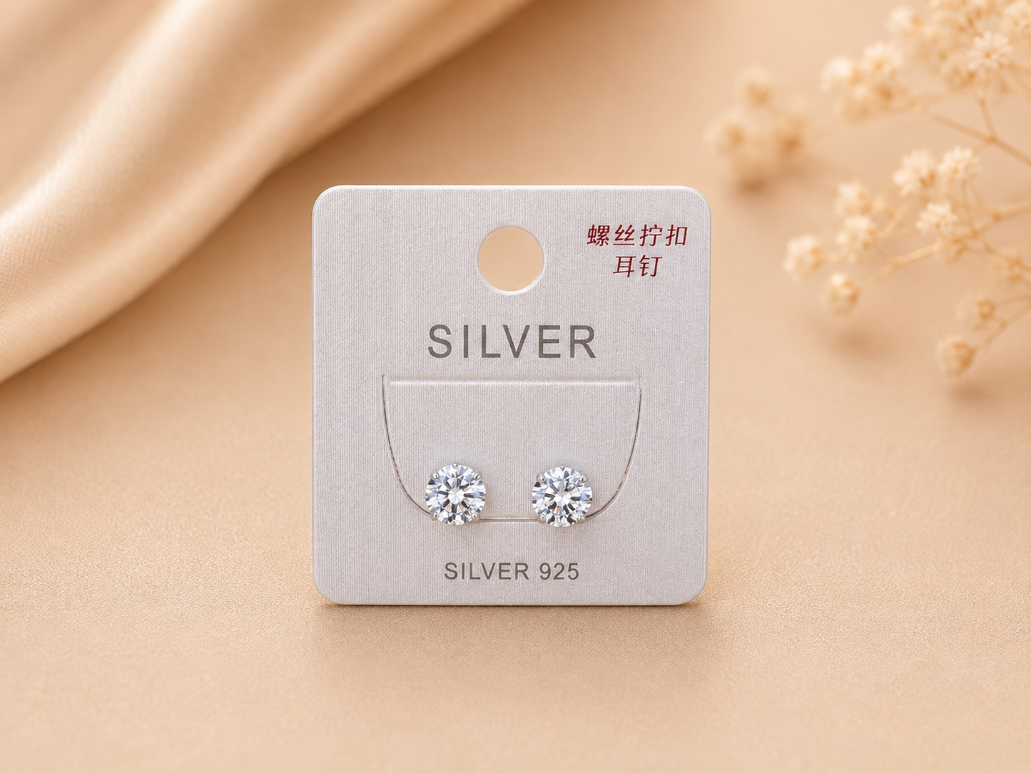 A pair of silver stud earrings displayed on a silver card labeled 'SILVER' with 'SILVER 925' below, set against a warm beige background with soft fabric and dried flowers.