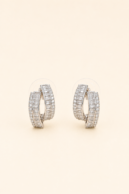 Silver crossover hoop earrings featuring baguette-cut and pavé crystals on a soft ivory cream background.