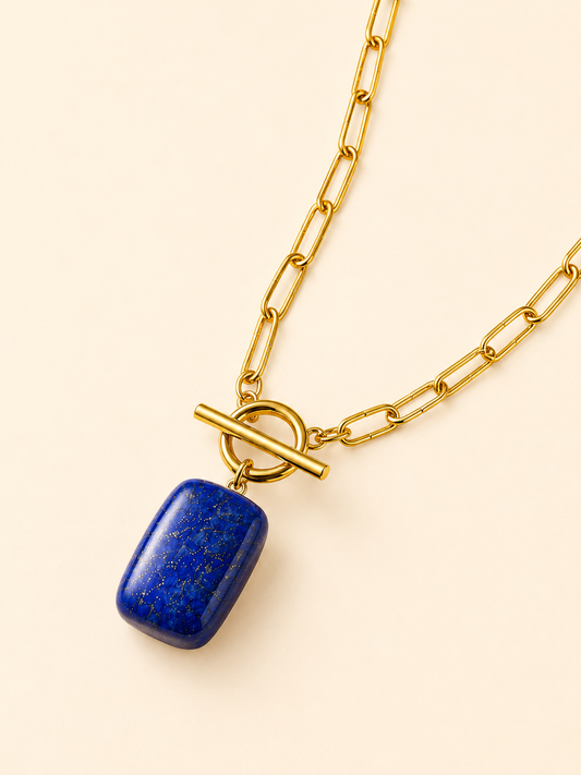 A gold chain necklace with a rectangular blue lapis lazuli pendant, featuring a toggle clasp closure, displayed on a light cream background.