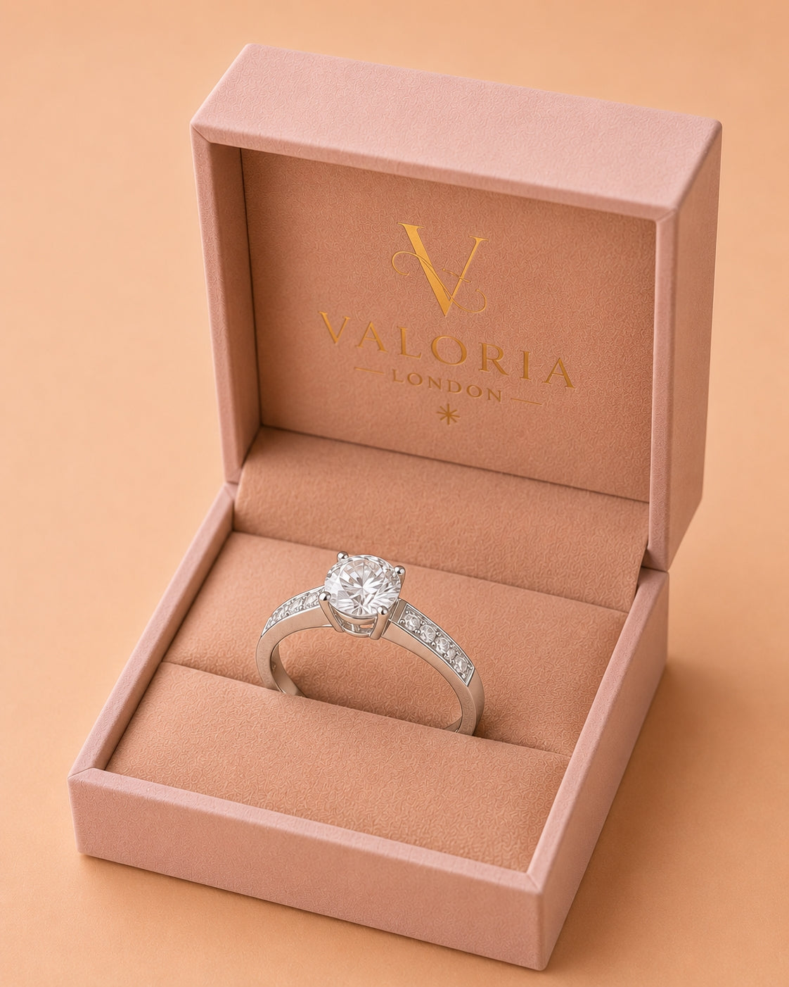 A silver solitaire engagement ring with a round-cut center stone and pavé-set side stones, displayed inside an open pink Valoria London jewelry box on a warm peach background.