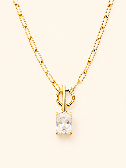 A gold paperclip chain necklace with a toggle clasp and a rectangular clear gemstone pendant, shown on a light cream background.