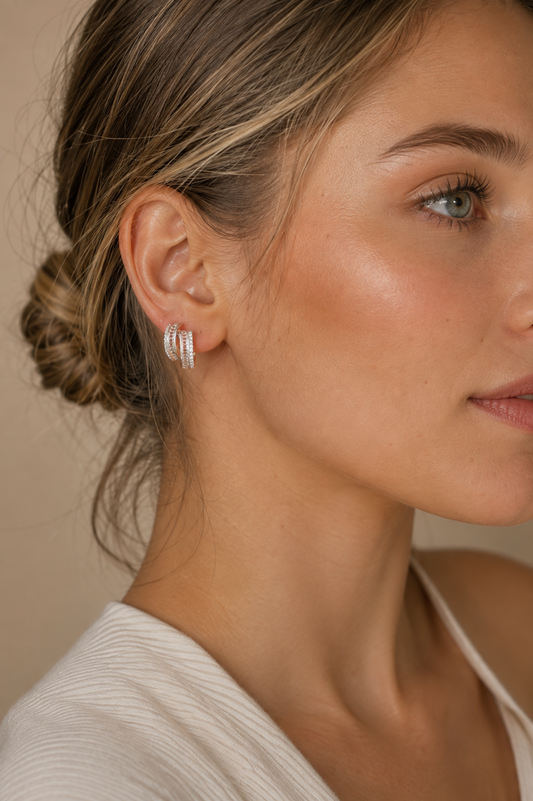 A close-up of a woman wearing a pair of silver hoop earrings adorned with small clear stones, set against a soft beige background.