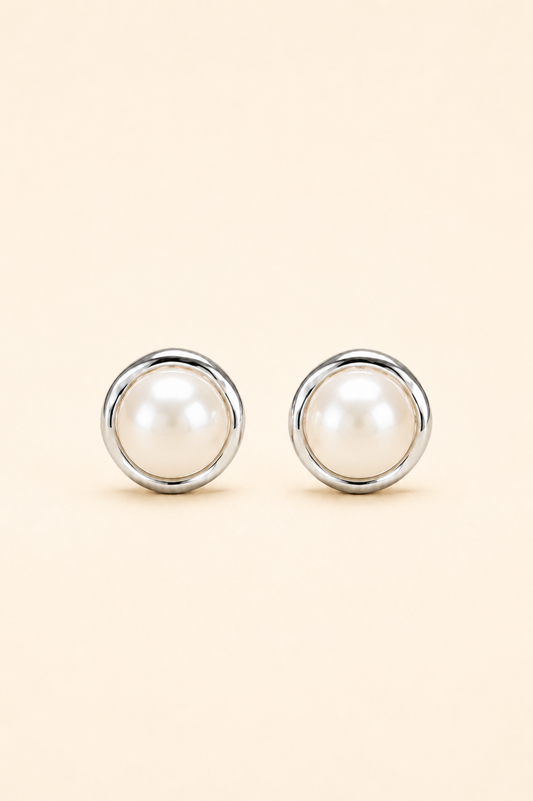 A pair of round stud earrings featuring white faux pearls set in polished silver-tone metal, displayed on a solid light beige background.