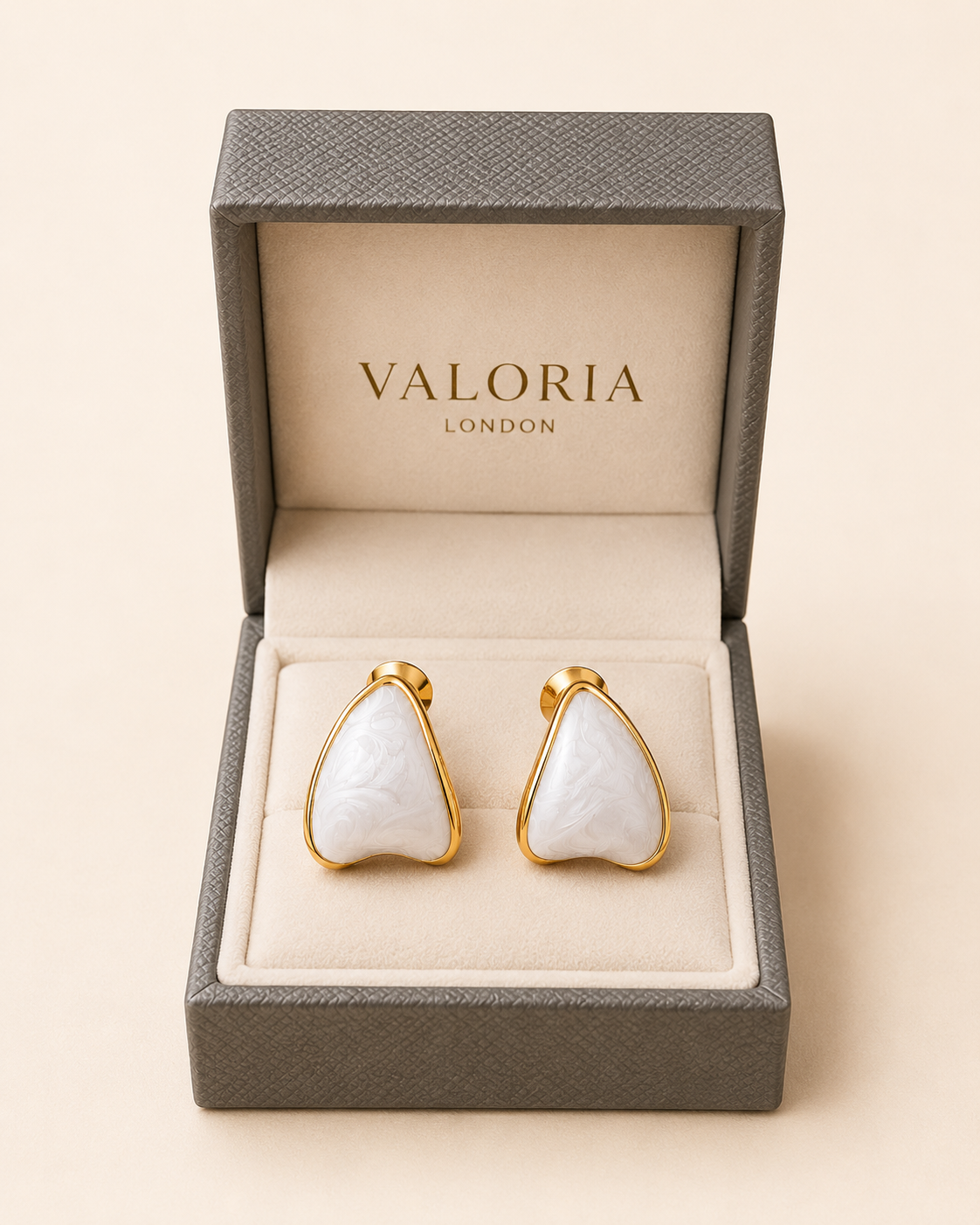 A pair of gold-tone stud earrings with white teardrop-shaped mother-of-pearl inlays, presented in an open textured gray Valoria London gift box on a light beige background.
