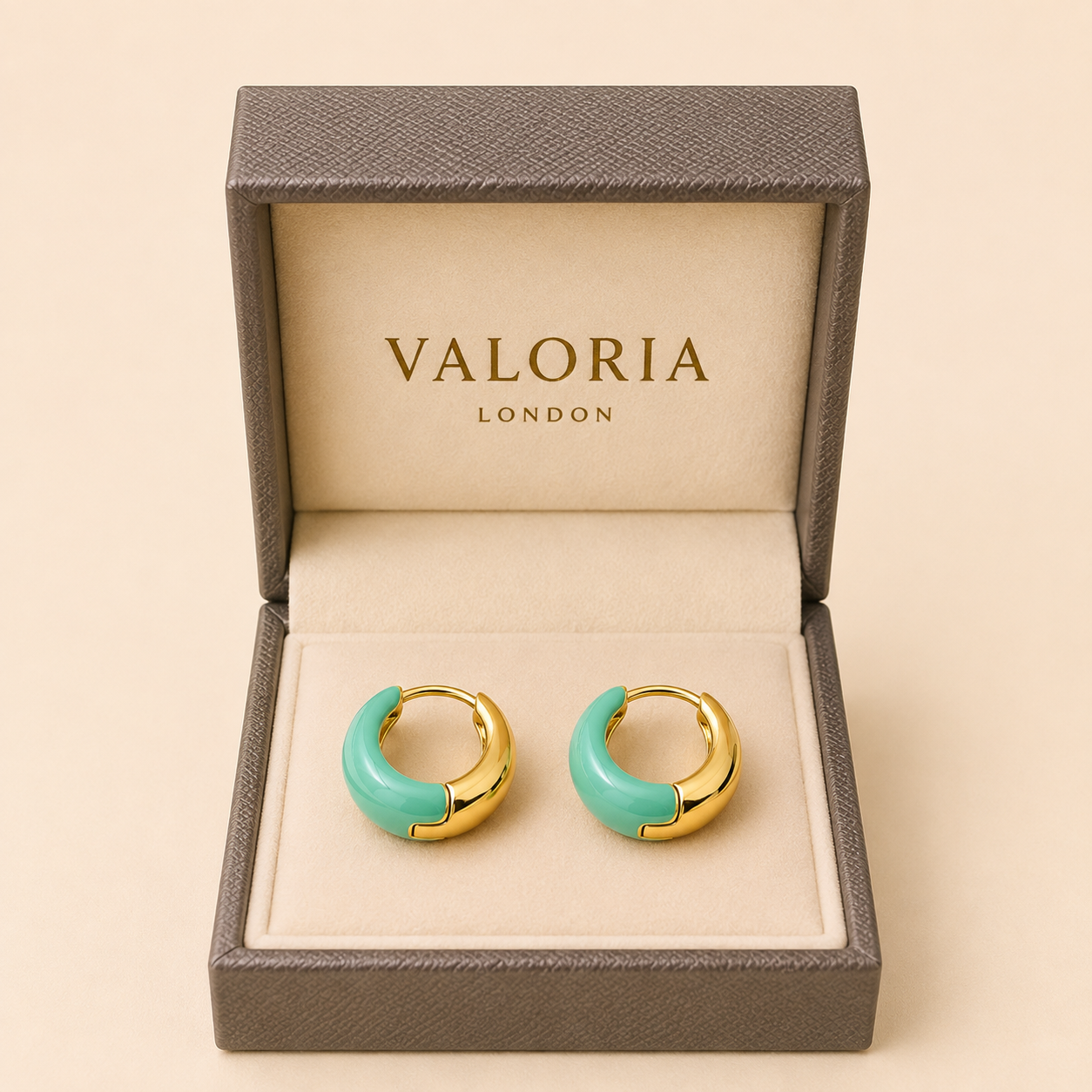 A pair of gold crescent hoop earrings with mint green enamel inlays, presented in an open textured gray Valoria London jewelry box on a beige background.