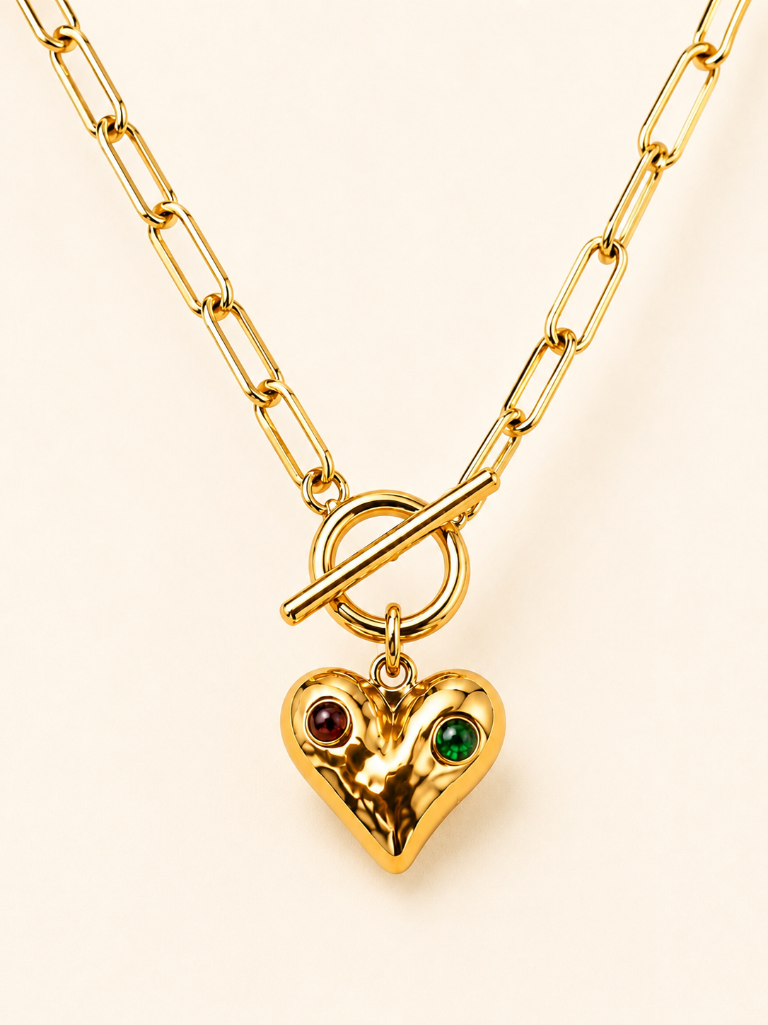 Gold paperclip chain necklace with a hammered heart pendant, red and green gemstone accents and toggle clasp on a soft ivory cream background