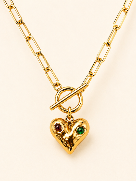 Gold paperclip chain necklace with a hammered heart pendant, red and green gemstone accents and toggle clasp on a soft ivory cream background