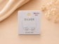 A pair of silver stud earrings displayed on a silver card labeled 'SILVER' with 'SILVER 925' below, set against a warm beige background with soft fabric and dried flowers.