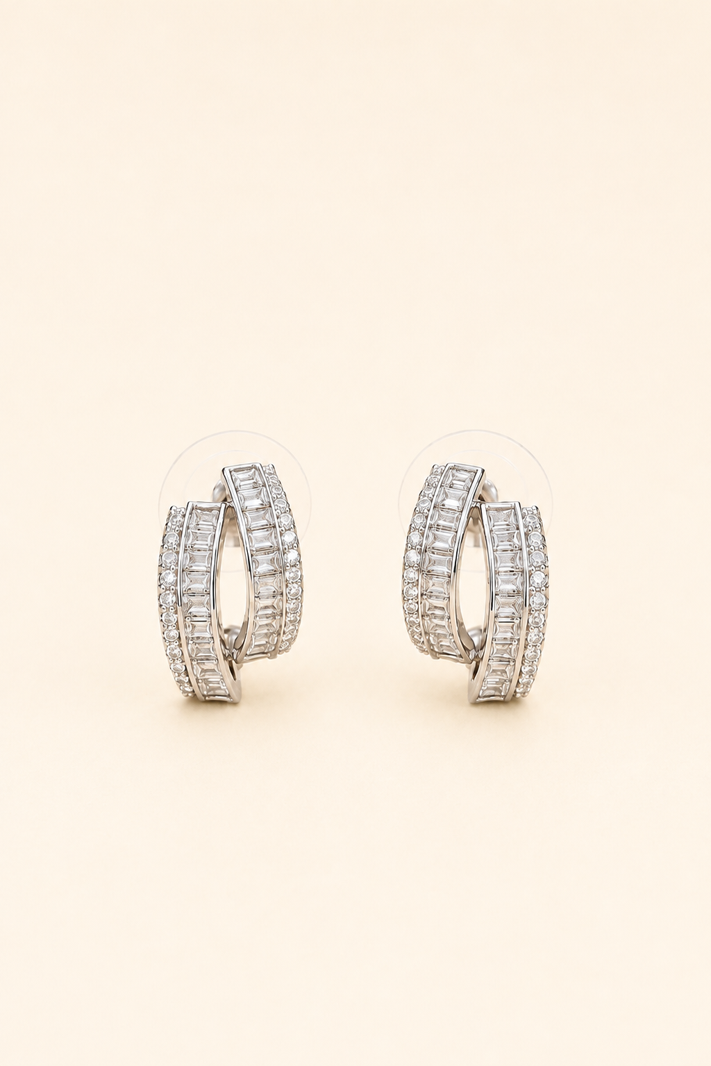 Silver crossover hoop earrings featuring baguette-cut and pavé crystals on a soft ivory cream background.
