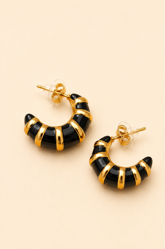 A pair of crescent-shaped hoop earrings with a glossy black enamel body and polished gold-tone metal bands, displayed on a solid light beige background.