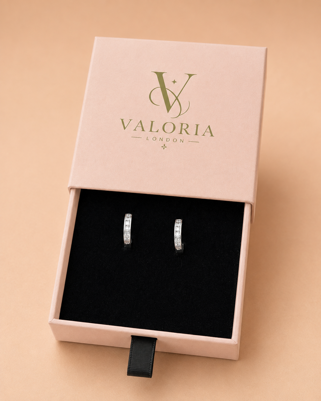 A pair of silver hoop earrings with clear stones, presented in a black-lined pink Valoria London gift box on a warm beige background.