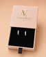 A pair of silver hoop earrings with clear stones, presented in a black-lined pink Valoria London gift box on a warm beige background.