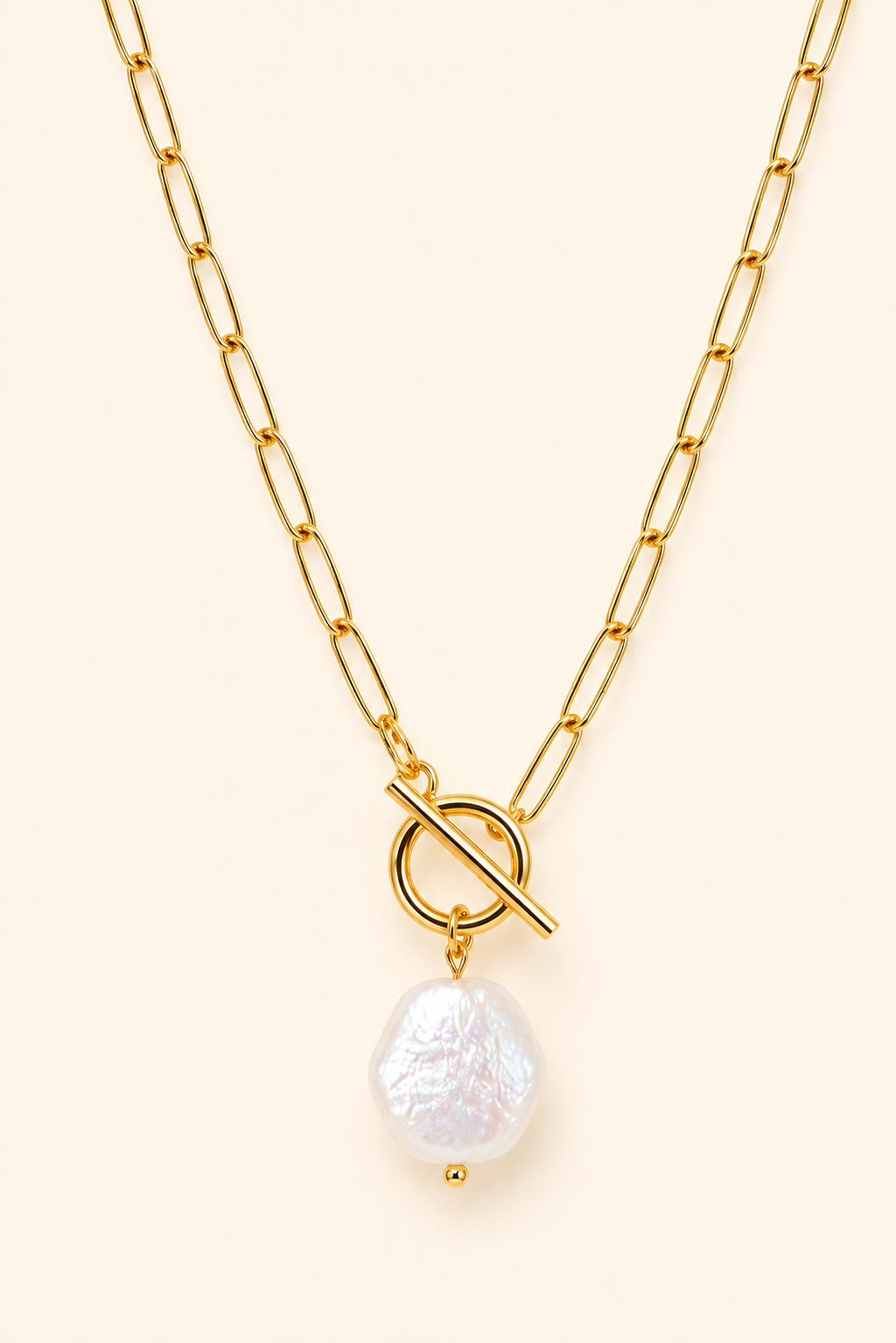 A gold chain necklace featuring a toggle clasp and a dangling baroque pearl pendant, displayed on a solid cream background.