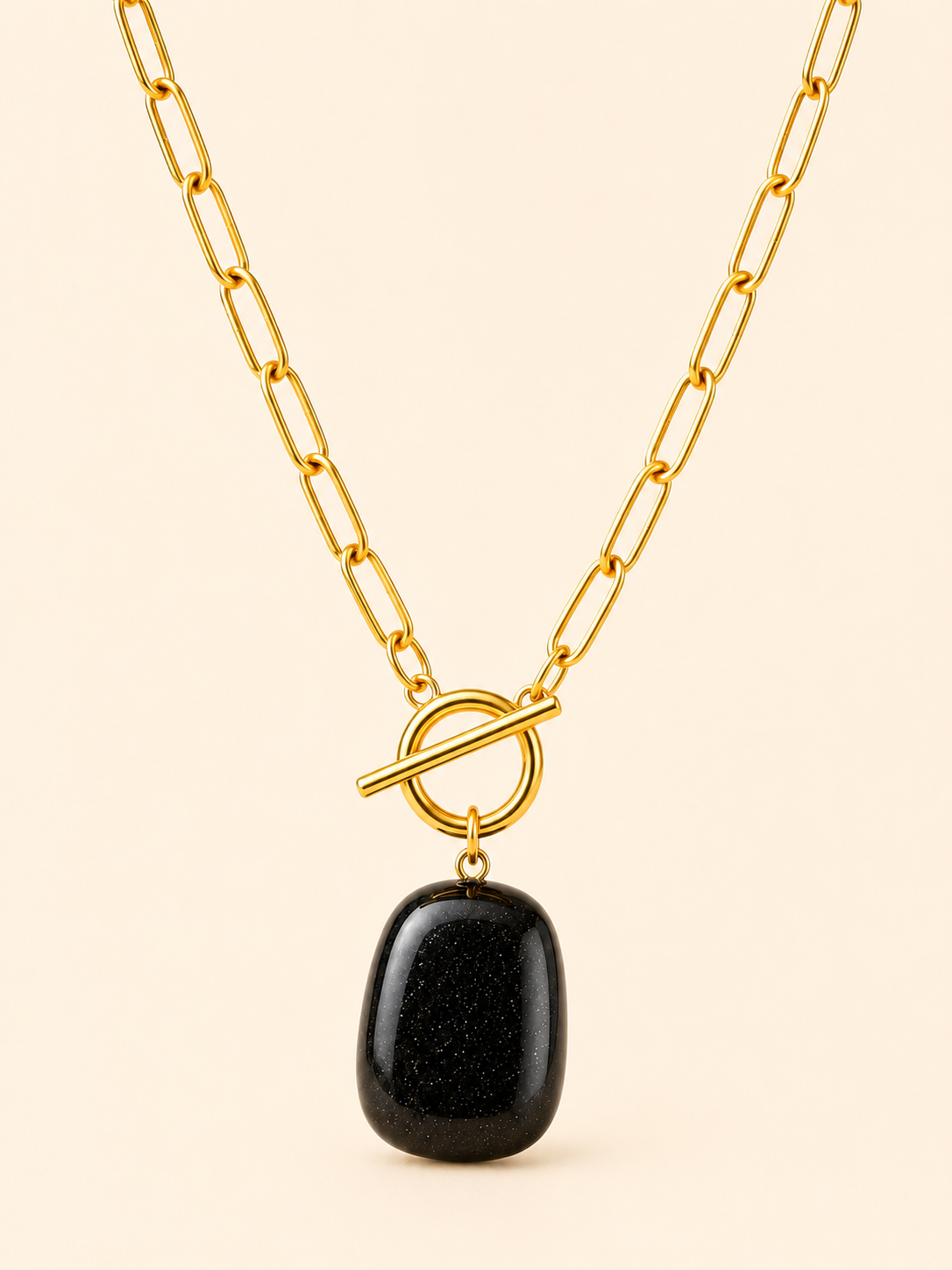 A gold paperclip chain necklace with a toggle clasp and a polished black stone pendant, shown against a solid light beige background.