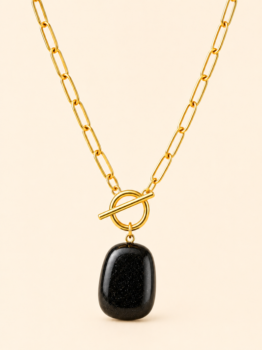 A gold paperclip chain necklace with a toggle clasp and a polished black stone pendant, shown against a solid light beige background.