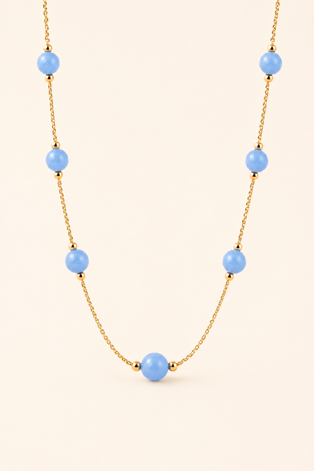A gold chain necklace featuring seven light blue round beads spaced along the chain, displayed against a solid pale beige background.
