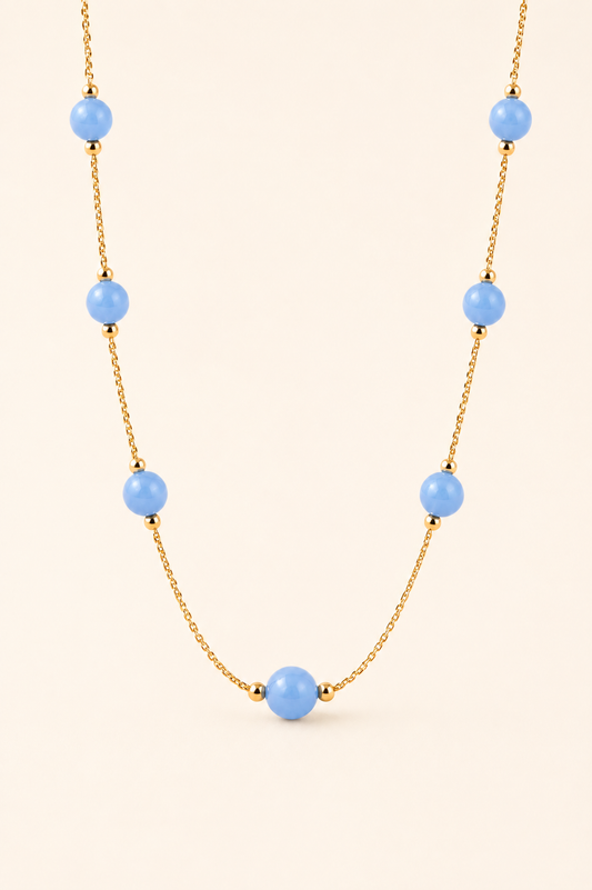A gold chain necklace featuring seven light blue round beads spaced along the chain, displayed against a solid pale beige background.