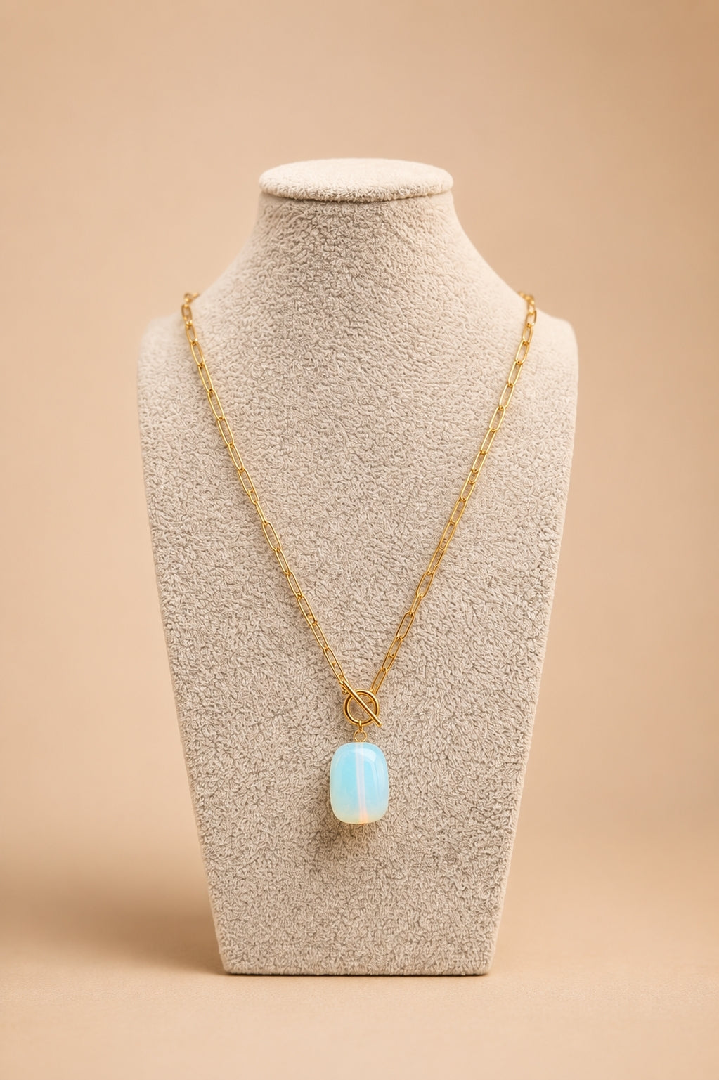 A gold chain necklace with a light blue rectangular pendant displayed on a textured beige jewelry bust against a warm beige background.
