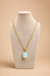 A gold chain necklace with a light blue rectangular pendant displayed on a textured beige jewelry bust against a warm beige background.