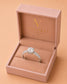 A silver solitaire engagement ring with a round-cut center stone and pavé-set side stones, displayed inside an open pink Valoria London jewelry box on a warm peach background.