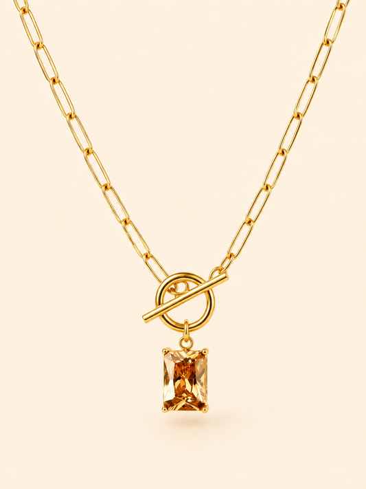 A gold paperclip chain necklace with a toggle clasp and a rectangular amber gemstone pendant, shown against a solid pale peach background.