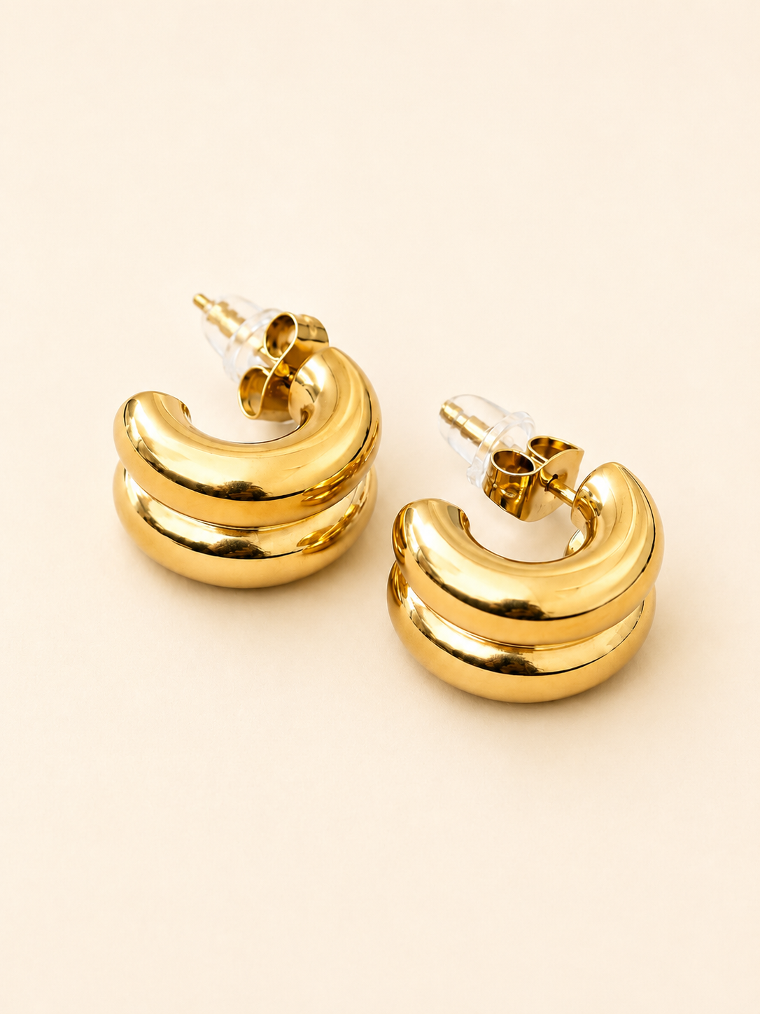 A pair of gold crescent-shaped hoop earrings with a polished finish and clear plastic backing, displayed on a light beige background.