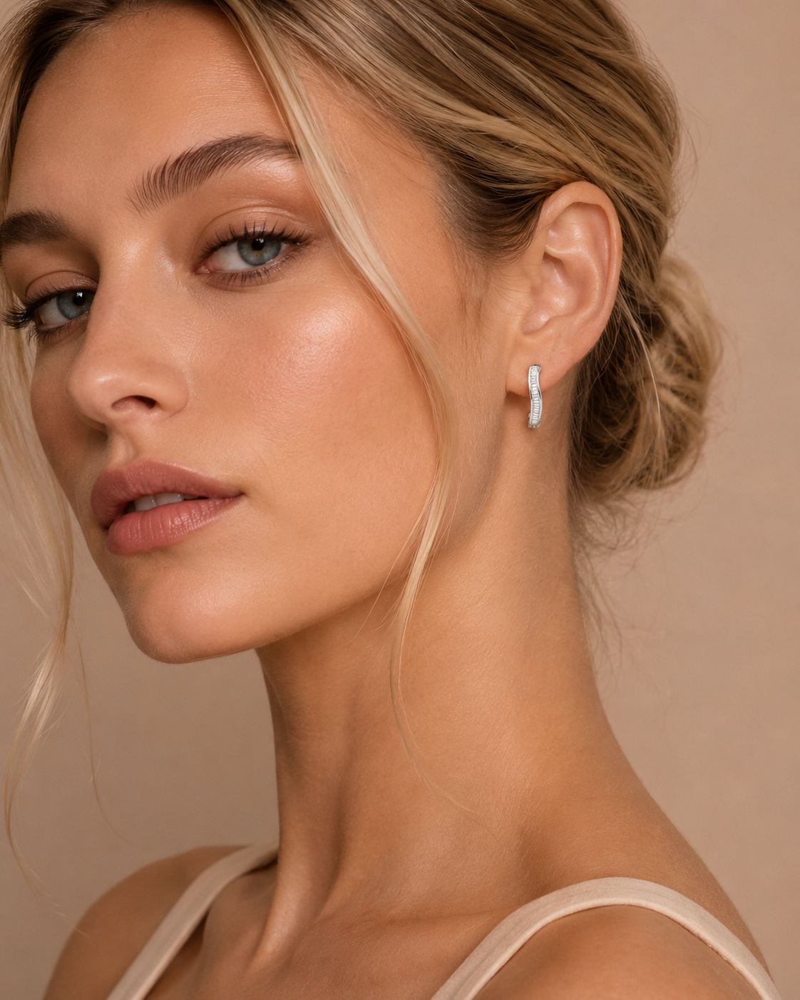 A close-up of a woman wearing a delicate silver crescent-shaped drop earring with a small white stone, set against a warm beige background.