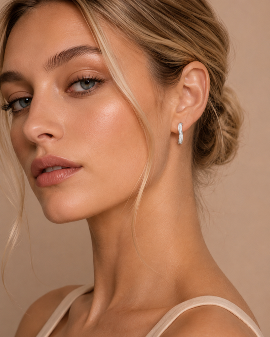 A close-up of a woman wearing a delicate silver crescent-shaped drop earring with a small white stone, set against a warm beige background.