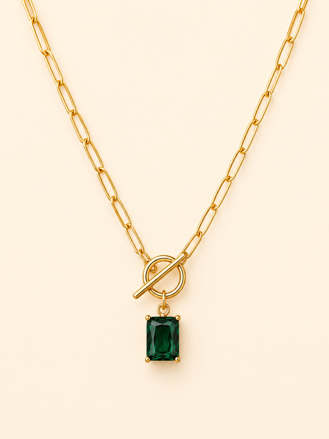 A gold paperclip chain necklace with a toggle clasp and a rectangular emerald-cut green gemstone pendant, displayed on a light cream background.