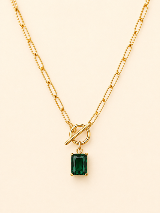 A gold paperclip chain necklace with a toggle clasp and a rectangular emerald-cut green gemstone pendant, displayed on a light cream background.