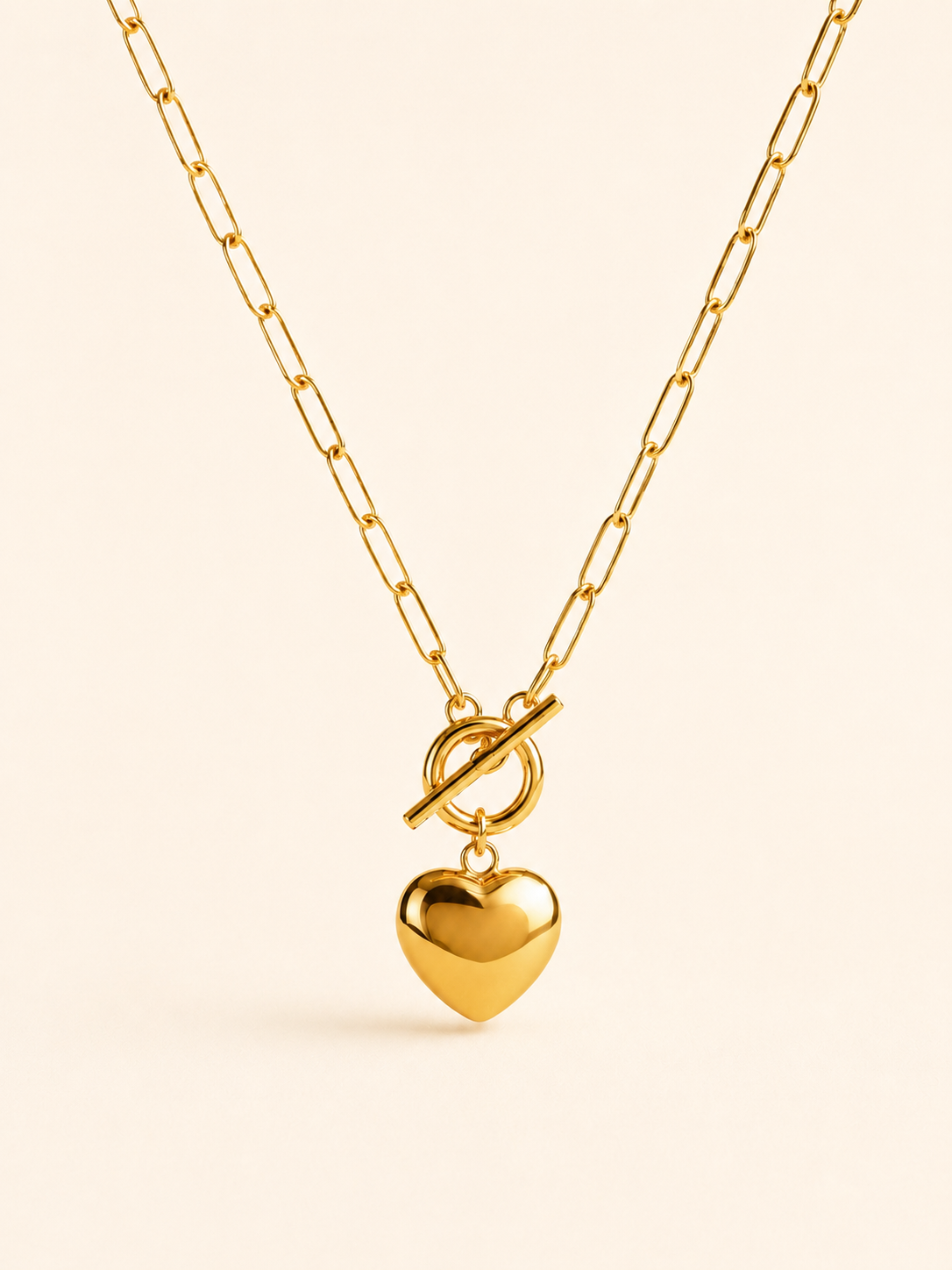 A gold paperclip chain necklace with a toggle clasp and a dangling heart-shaped charm, shown on a light cream background.