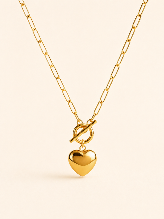 A gold paperclip chain necklace with a toggle clasp and a dangling heart-shaped charm, shown on a light cream background.