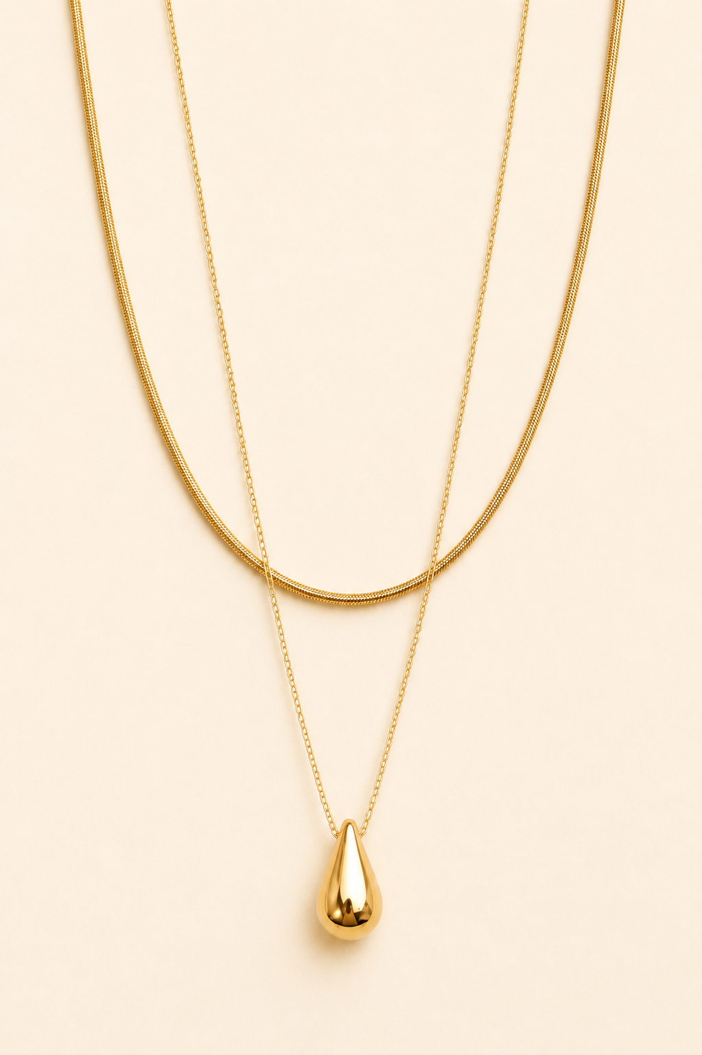 A double-layered gold necklace set featuring a thick snake chain and a delicate chain with a polished teardrop pendant, displayed on a solid light beige background.