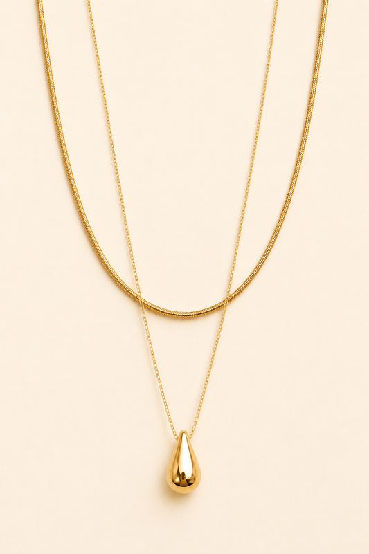 A double-layered gold necklace set featuring a thick snake chain and a delicate chain with a polished teardrop pendant, displayed on a solid light beige background.