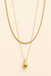 A double-layered gold necklace set featuring a thick snake chain and a delicate chain with a polished teardrop pendant, displayed on a solid light beige background.