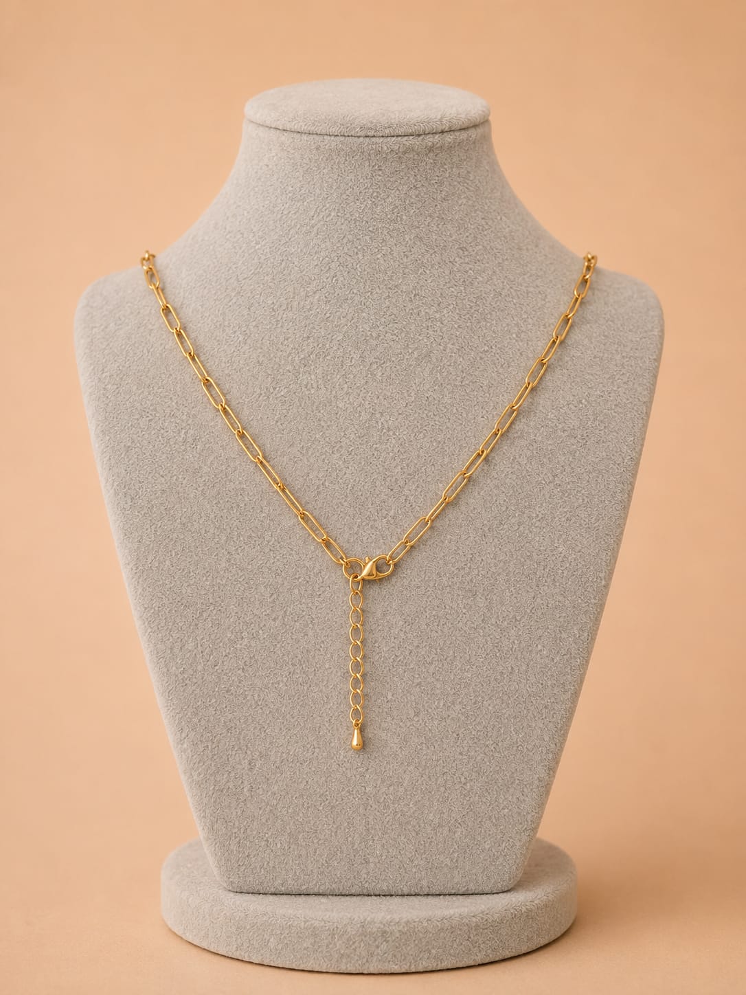 A gold chain necklace with a small clasp and a dangling chain extender, displayed on a light gray jewelry bust against a warm peach background.