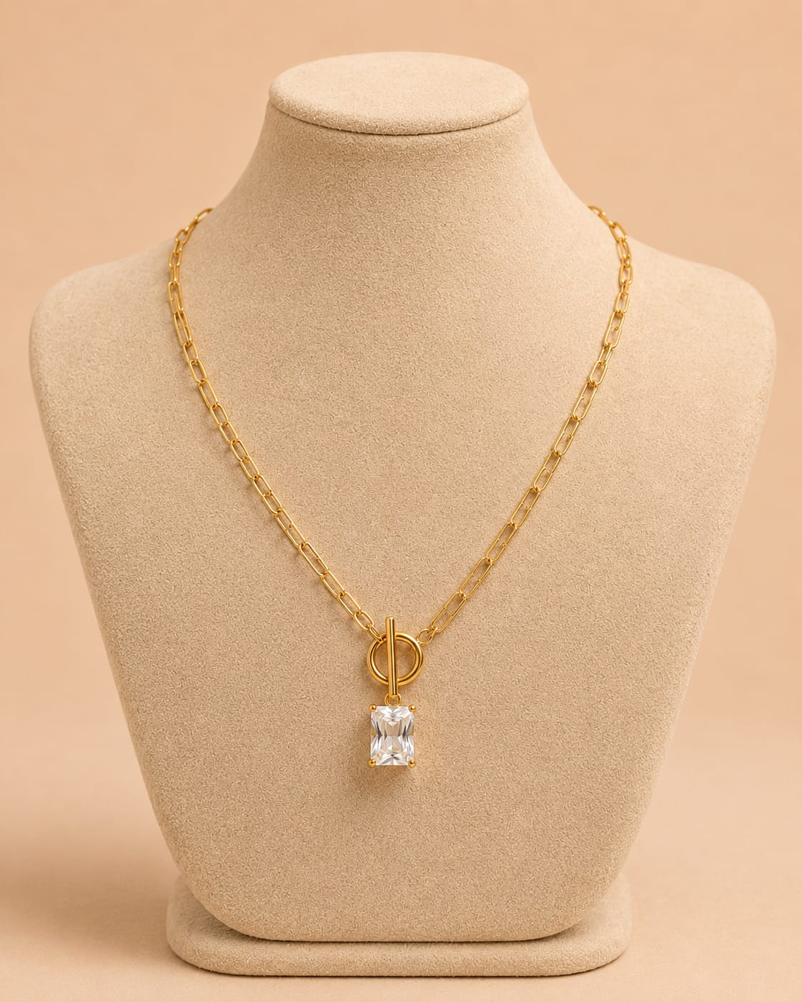 A gold chain necklace with a clear rectangular gemstone pendant, displayed on a beige jewelry bust against a warm beige background.