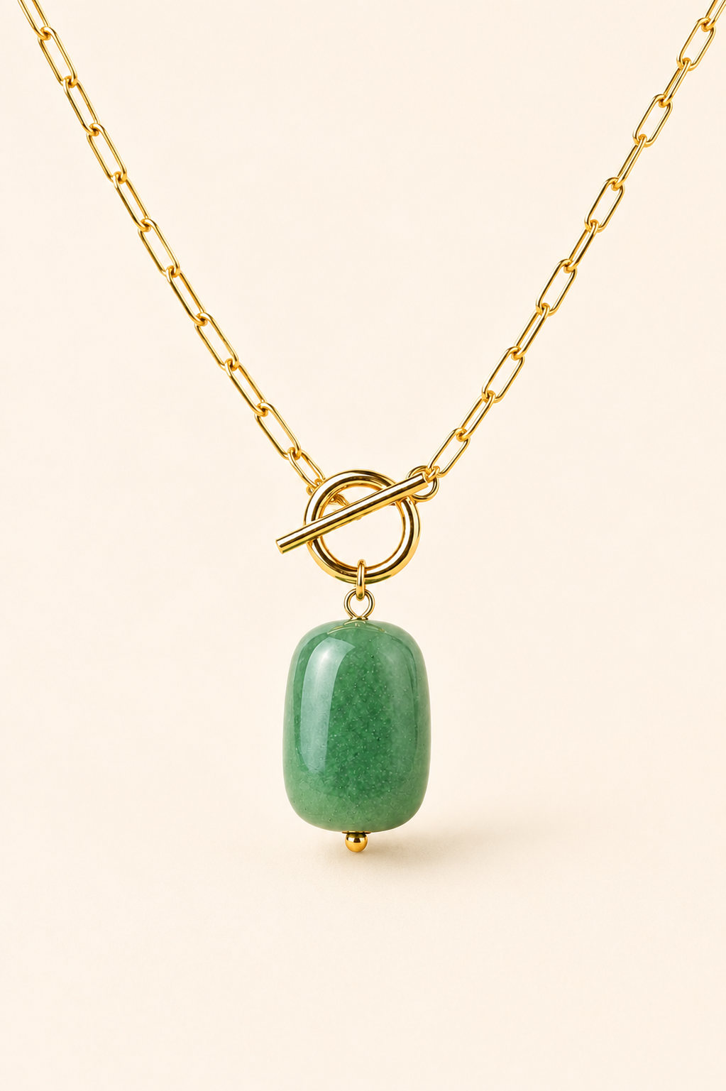 A gold chain necklace with a toggle clasp and a polished green stone pendant, shown against a solid light beige background.