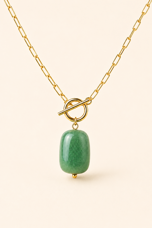 A gold chain necklace with a toggle clasp and a polished green stone pendant, shown against a solid light beige background.