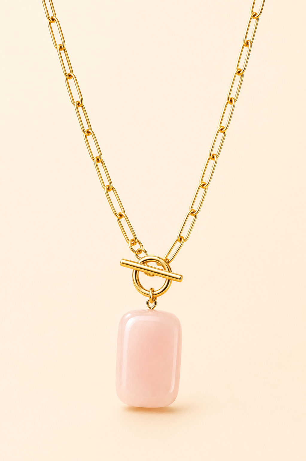 A gold paperclip chain necklace with a toggle clasp and a smooth, rectangular pale pink stone pendant, shown against a solid light beige background.