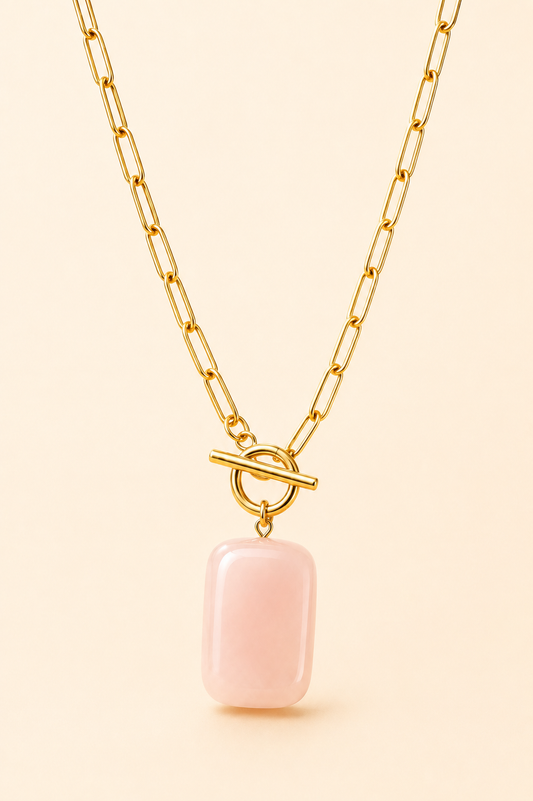 A gold paperclip chain necklace with a toggle clasp and a smooth, rectangular pale pink stone pendant, shown against a solid light beige background.