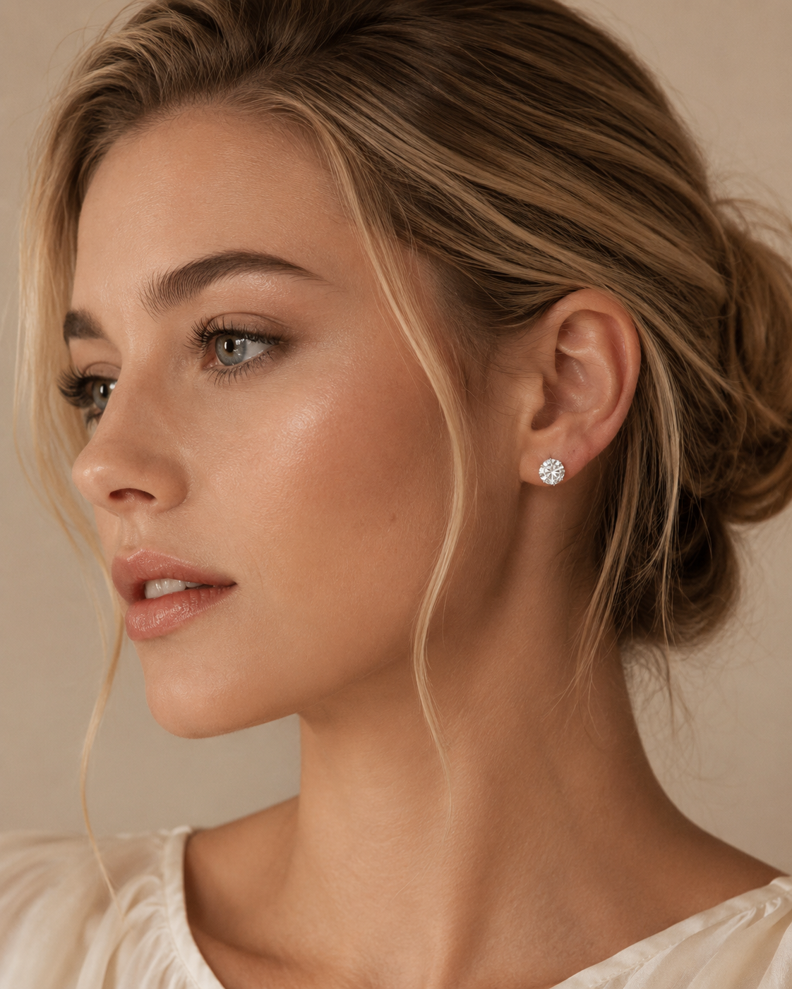 A close-up portrait of a woman with blonde hair in an updo, wearing a small round brilliant-cut diamond stud earring. The background is a soft, warm beige studio backdrop.