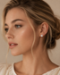 A close-up portrait of a woman with blonde hair in an updo, wearing a small round brilliant-cut diamond stud earring. The background is a soft, warm beige studio backdrop.