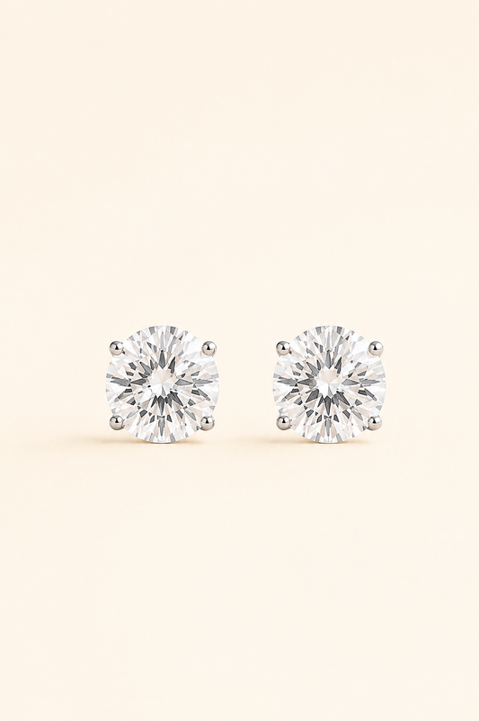 A pair of round-cut stud earrings with clear gemstones in a silver-toned prong setting, displayed on a light cream background.