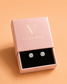 Round solitaire crystal stud earrings presented in a pink Valoria London jewellery box on a warm nude peach background.