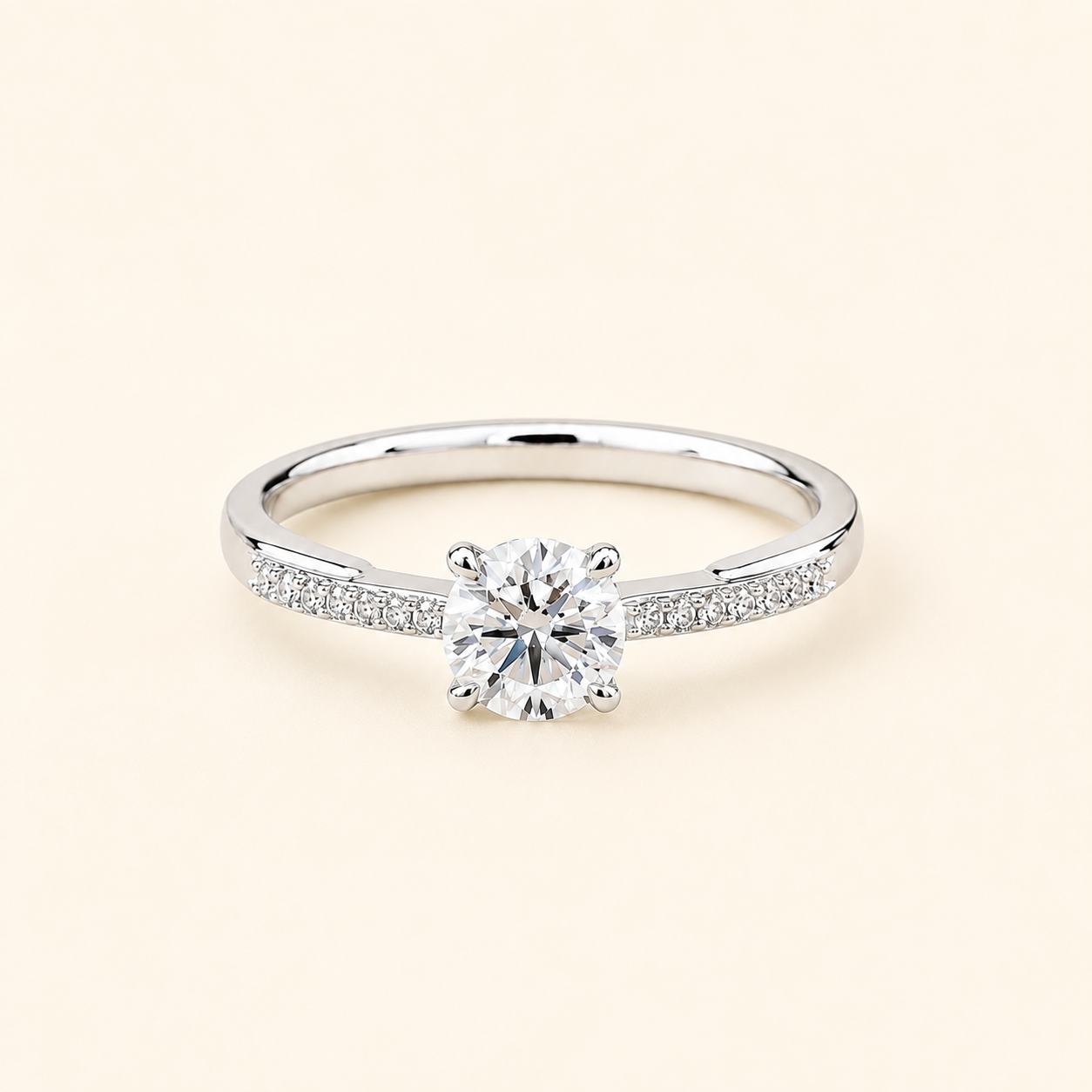 A silver-tone solitaire engagement ring with a round-cut center stone and pavé-set accent stones along the band, shown on a light cream background.