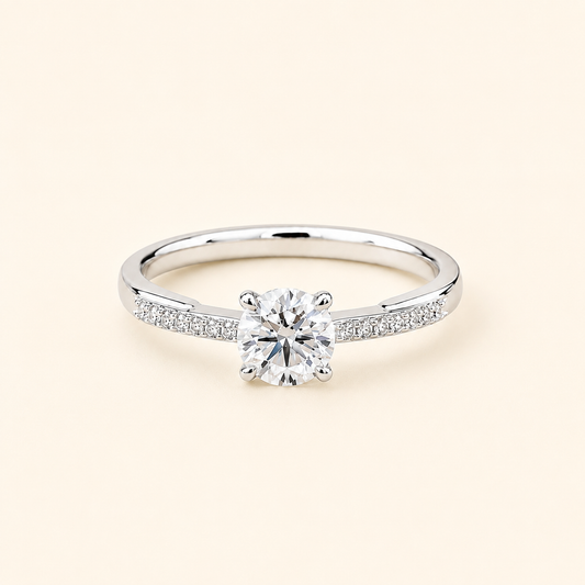 A silver-tone solitaire engagement ring with a round-cut center stone and pavé-set accent stones along the band, shown on a light cream background.