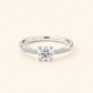A silver-tone solitaire engagement ring with a round-cut center stone and pavé-set accent stones along the band, shown on a light cream background.