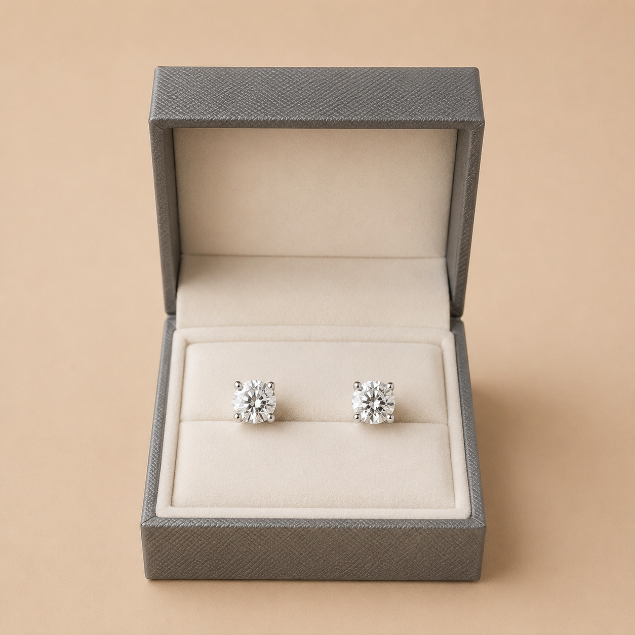 A pair of round-cut diamond stud earrings displayed in an open textured gray jewelry box with a beige velvet insert, set against a solid beige background.