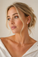 A close-up portrait of a woman with blonde hair in a low bun, wearing a delicate gold chain necklace and a small round stud earring with a clear gemstone, set against a soft beige background.